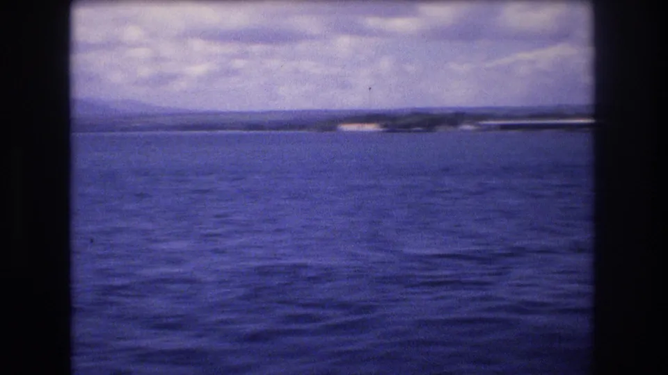 Final 1977 Honolulu-area coastal frame with rippled blue water, a low far shoreline, bright light on the distant coast, and cloudy sky.