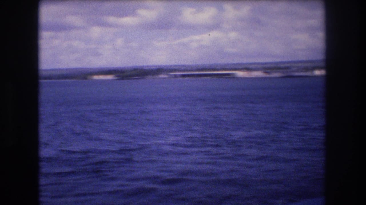 1977 8mm home-movie frame from the Honolulu, Hawaii metadata showing open water, a low distant shoreline, and clouds under a blue-purple film cast.
