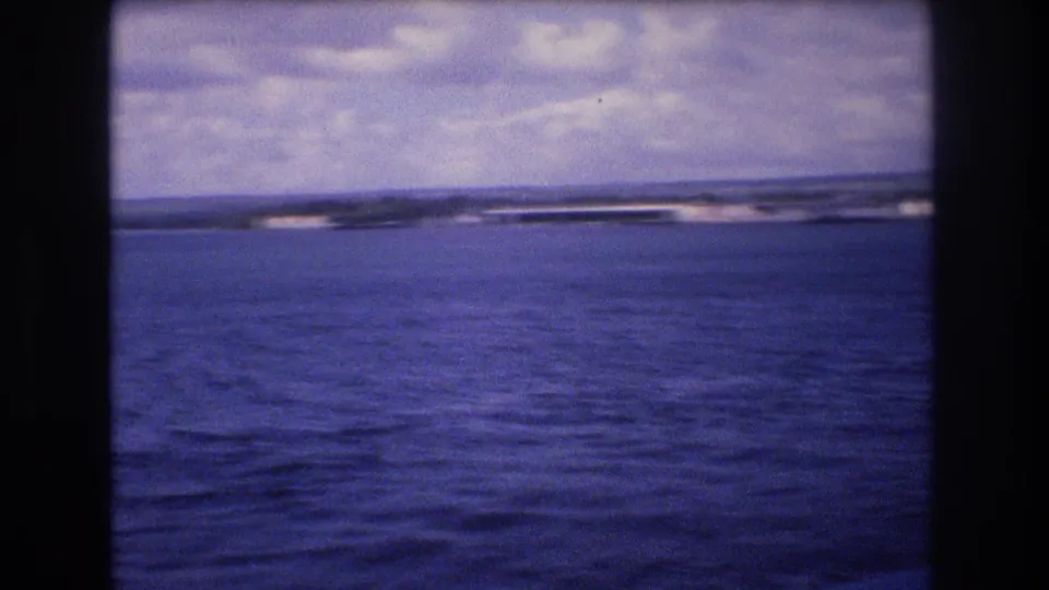 1977 8mm home-movie frame from the Honolulu, Hawaii metadata showing open water, a low distant shoreline, and clouds under a blue-purple film cast.