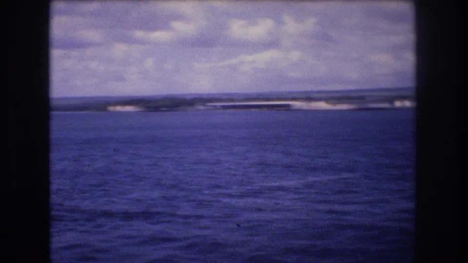 The frame continues the boat-level pan across blue water toward a low shoreline, with soft focus and aged-film color typical of the 8mm source.