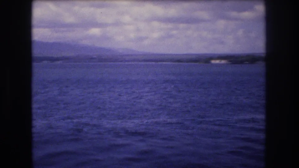 Final frame of the 1977 Honolulu-labeled 8mm clip shows open water, hazy hills, distant shoreline structures, and a dark foreground edge that may relate to the boat or a nearby figure.