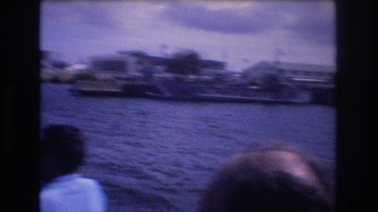 1977 Honolulu 8mm frame looking from a moving boat across blue water toward a soft, distant shoreline, with passengers partially visible at the bottom edge.