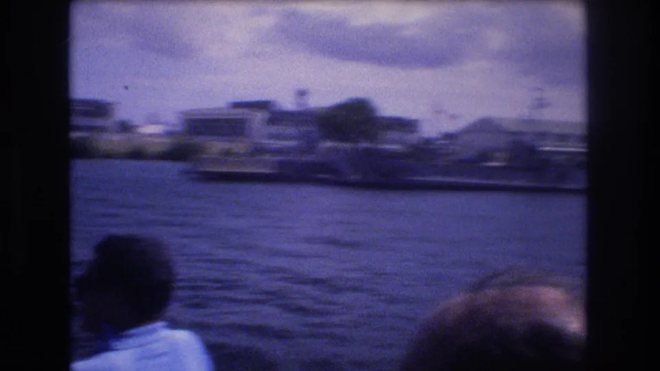 1977 Honolulu home-movie frame from a boat, showing a seated passenger from behind, rippled water, and blurred waterfront buildings under a cloudy sky.