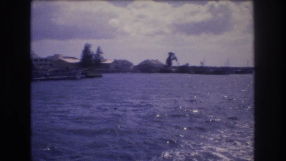 1977 Honolulu boat-view frame with open water, sun glints on small waves, a distant shoreline, trees, and low harbor structures beneath a cloudy sky.