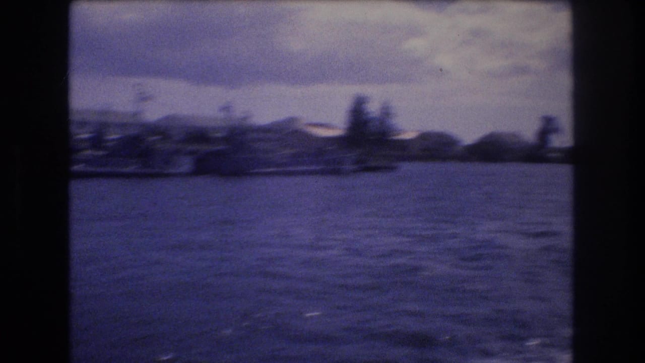 1977 Honolulu home-movie frame showing choppy harbor water, a distant low-rise shoreline, palm shapes, cloudy sky, and dark film gate edges.