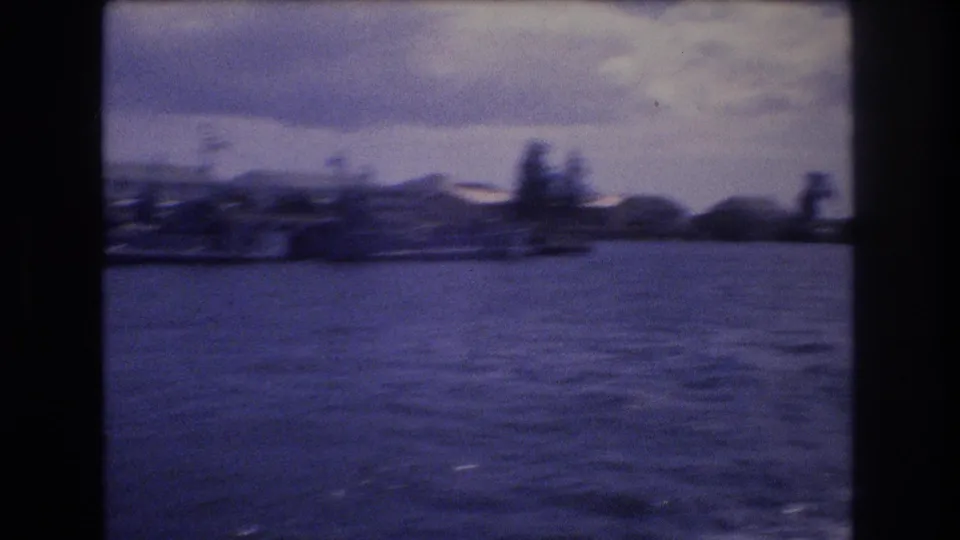 1977 Honolulu home-movie frame showing choppy harbor water, a distant low-rise shoreline, palm shapes, cloudy sky, and dark film gate edges.