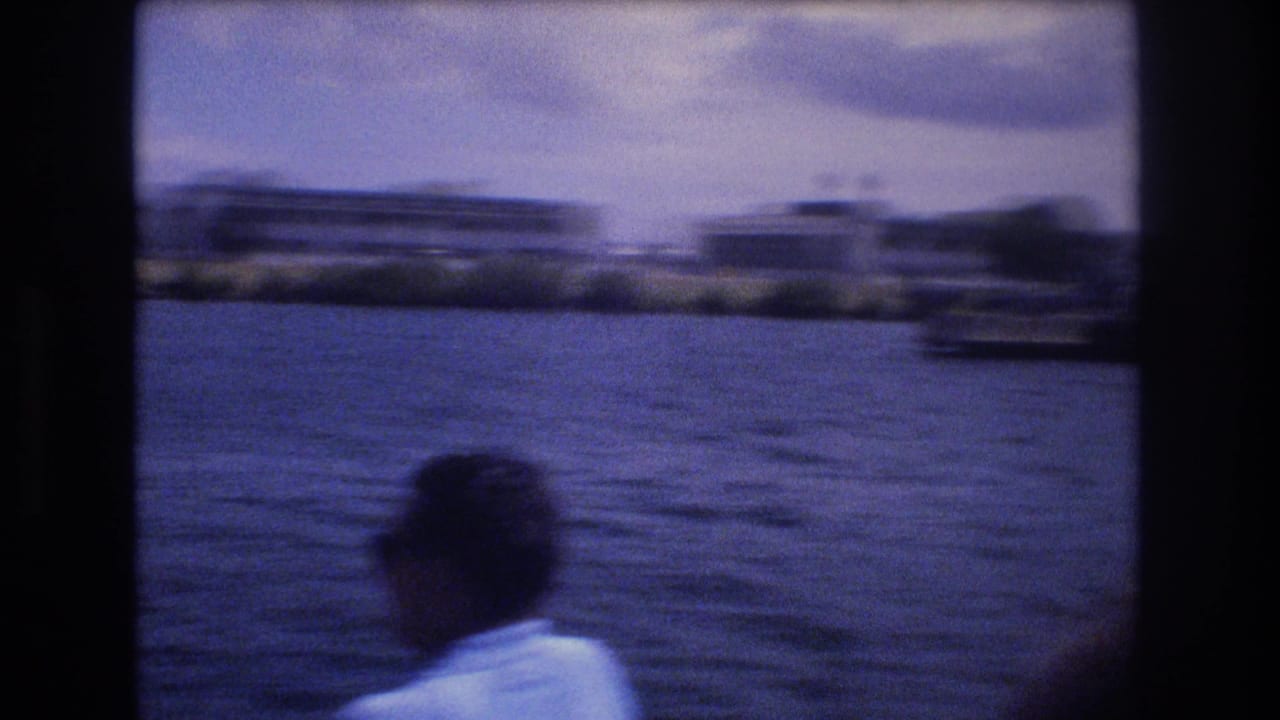 1977 Honolulu boat-view frame with a foreground person in a light shirt looking toward low-rise waterfront buildings across choppy water.