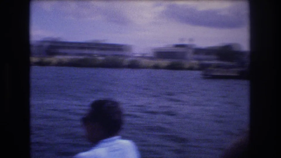 1977 Honolulu boat-view frame with a foreground person in a light shirt looking toward low-rise waterfront buildings across choppy water.