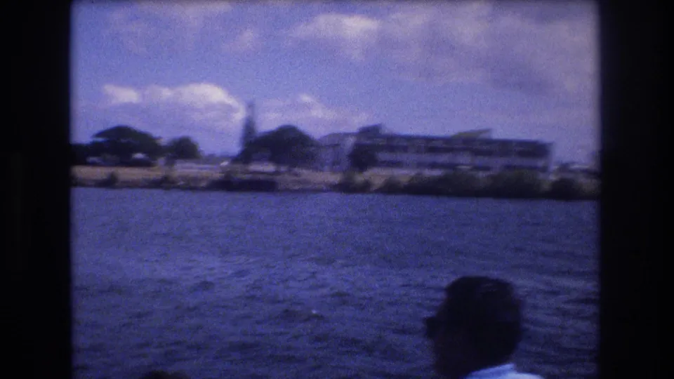 1977 Honolulu home-movie frame showing waterfront buildings, palms, blue harbor water, a partly cloudy sky, and a partially visible passenger at the lower edge.