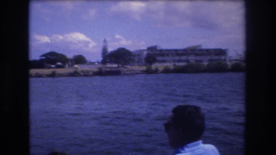 1977 Honolulu boat-view frame with a foreground passenger, choppy water, palm-lined shore, low buildings, and soft 8mm color shift.