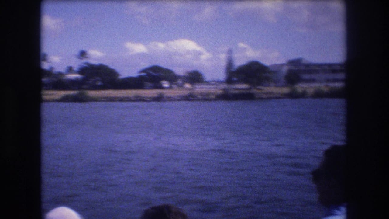 1977 Honolulu home-movie frame looking from inside a boat across blue water toward a low shoreline with trees and indistinct buildings.