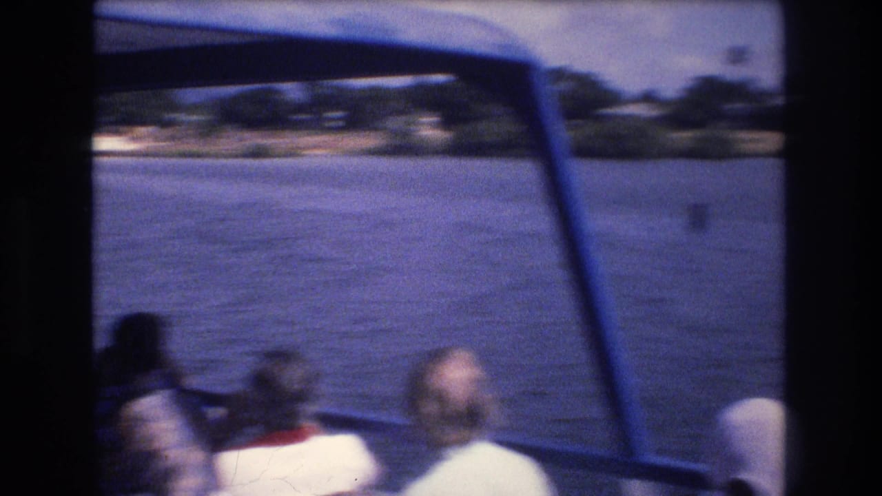 Tourists seated near the front of a boat in 1977 Honolulu look across the water toward a distant shoreline, with the boat canopy and window supports framing the view.