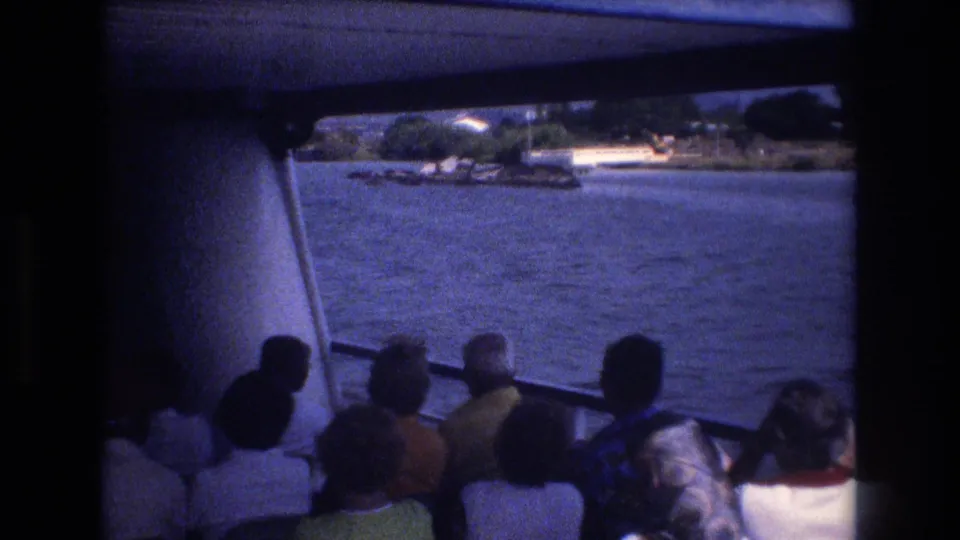 From behind a group of tourists on a Honolulu boat tour, the frame shows passengers facing the water with a shoreline, trees, and small waterfront structures in the distance.