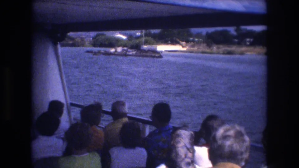 A 1977 Honolulu 8mm frame shows tourists seated beneath the boat canopy, viewed from behind as the vessel passes blue water and a low shoreline.