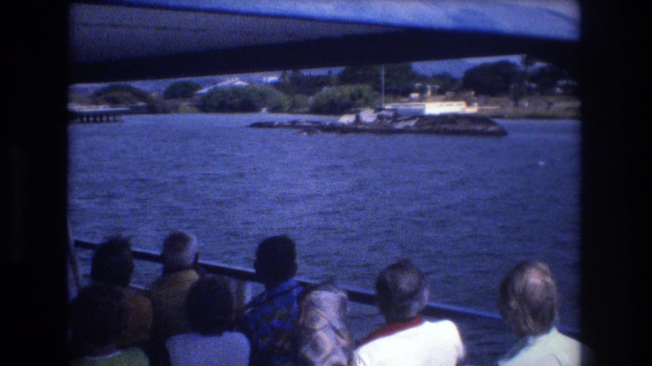 1977 Honolulu home-movie frame showing tourists seated under a boat canopy, facing open water and a distant shoreline with greenery and waterfront structures.