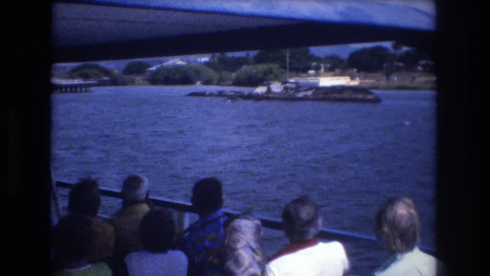 1977 Honolulu home-movie frame showing tourists seated under a boat canopy, facing open water and a distant shoreline with greenery and waterfront structures.