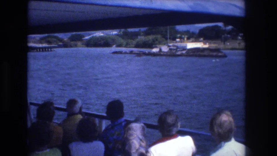 1977 Honolulu travel footage frame with seated passengers beneath the canopy, open blue water, and a distant shoreline visible beyond the boat railing.