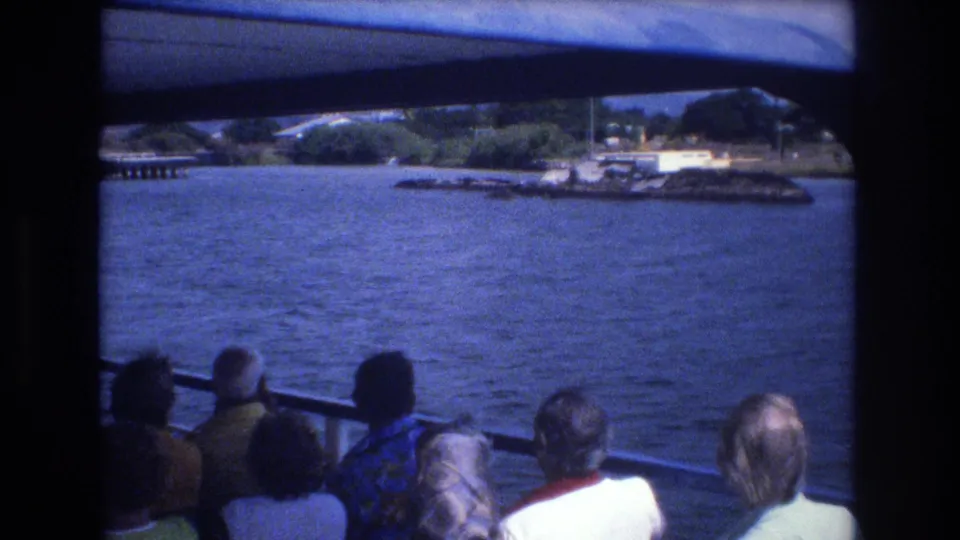 1977 Honolulu 8mm frame showing tourists in casual clothing seated along a boat railing as the camera looks over water toward a green shoreline and waterfront structures.