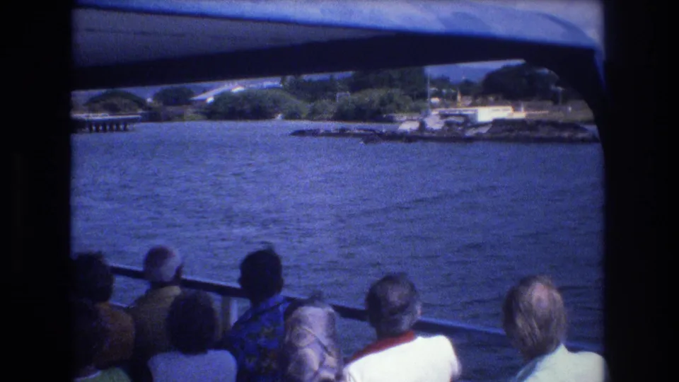 1977 Honolulu home-movie frame with passengers seen from behind beneath a canopy, looking across open water toward shoreline vegetation and waterfront features.