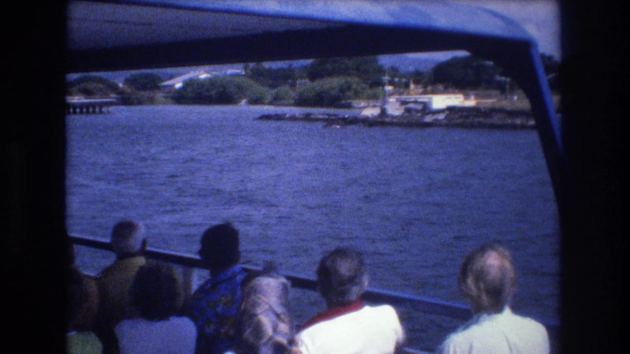 1977 Honolulu home-movie frame showing passengers from behind on a tour boat, with blue water, shoreline trees, and dock structures ahead.