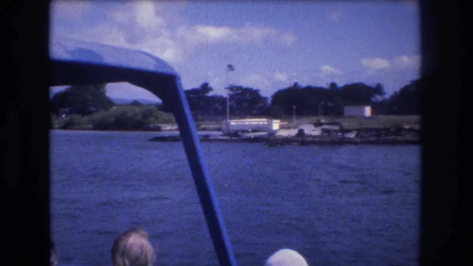 Honolulu boat-ride view with the tour boat frame in the foreground, a passenger partly visible, and a tree-lined shoreline with dock structures across the water.