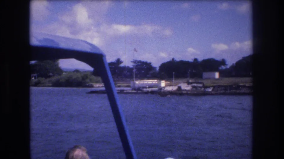 Final 1977 Honolulu tour-boat frame showing blue water, a distant shoreline with trees and dock structures, a flagpole, and a partial passenger at the bottom edge.