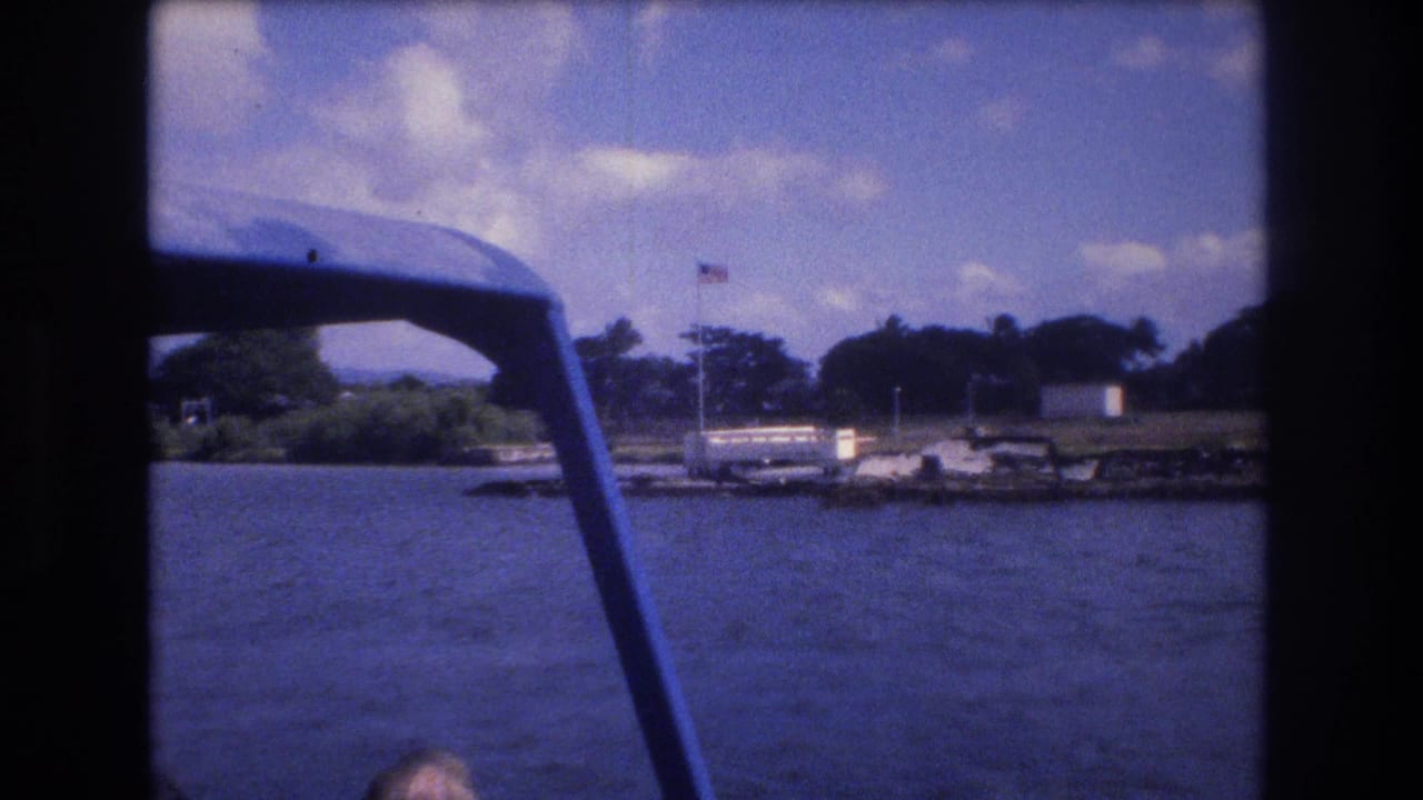 1977 8mm Honolulu boat-view frame with blue canopy support in the foreground, rippled water, a distant dock area, shoreline trees, and an American flag across the water.