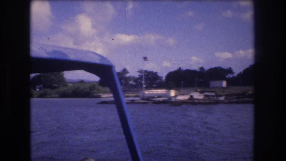 1977 8mm boat-view frame in Honolulu with a blue canopy support, choppy water, a distant dock and small shoreline structure, dense trees, and an American flag.