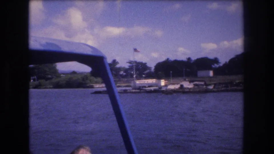 1977 Honolulu 8mm frame from a moving boat, with rippled water in front, blue canopy support at left, dock structures, a small white building, trees, and an American flag on shore.
