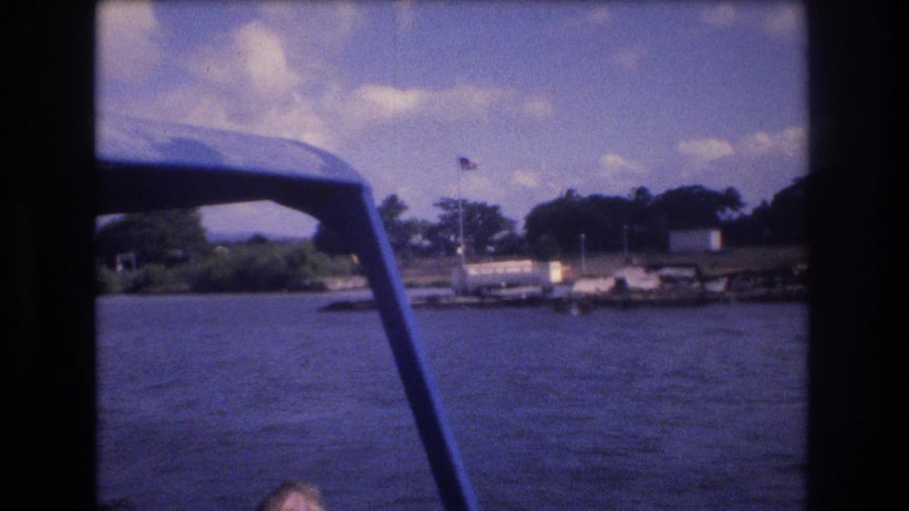 1977 Honolulu-area home-movie view from a moving boat, with a blue canopy frame crossing the foreground and an American flag near the waterfront shoreline.