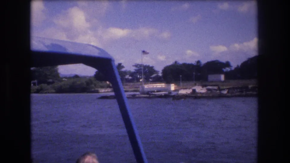 A moving-boat perspective in Honolulu, 1977, showing harbor water, a blue canopy frame, shoreline trees, low buildings, and an American flag on a pole.