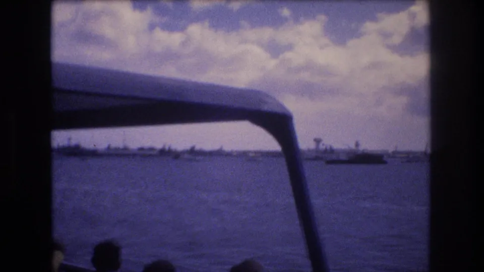 The 1977 Honolulu boat view looks across open harbor water toward distant craft and low shoreline, with the blue canopy frame and passenger silhouettes in the foreground.