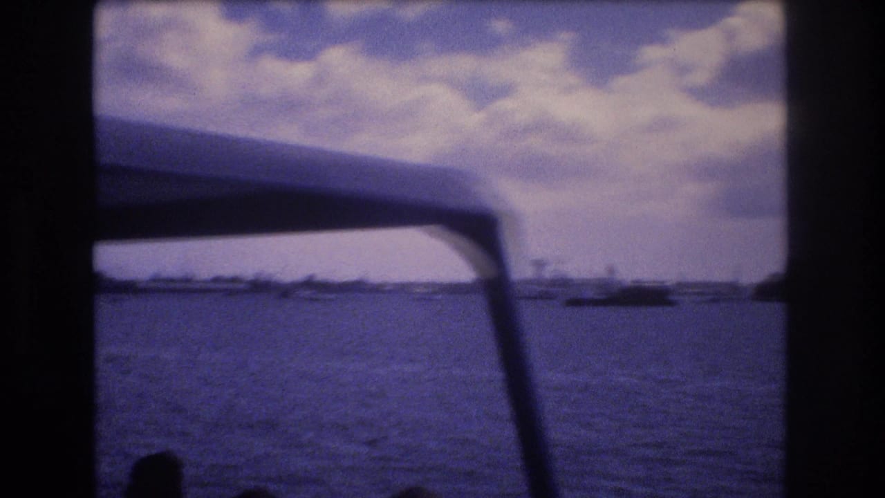 1977 Honolulu harbor view from a moving boat, with rippled water, distant boats, shoreline, clouds, and a dark curved canopy in the foreground.