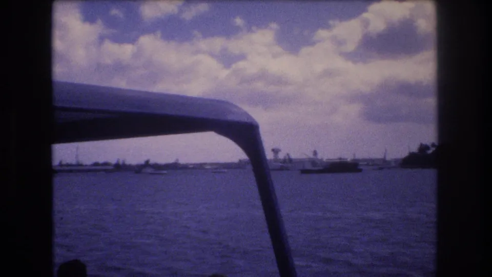 1977 Honolulu waterfront seen from a boat, with small vessels on calm water, a low shore, distant tower-like forms, and a dark canopy edge crossing the frame.
