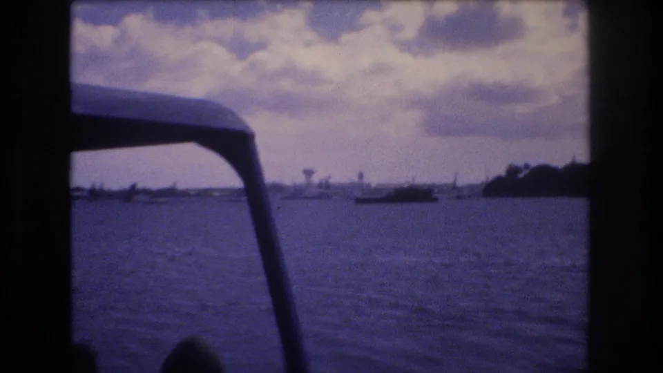 Vintage 8mm Honolulu harbor frame showing rippled blue water, a distant boat, low land, clouds, and the foreground edge of the boat canopy.