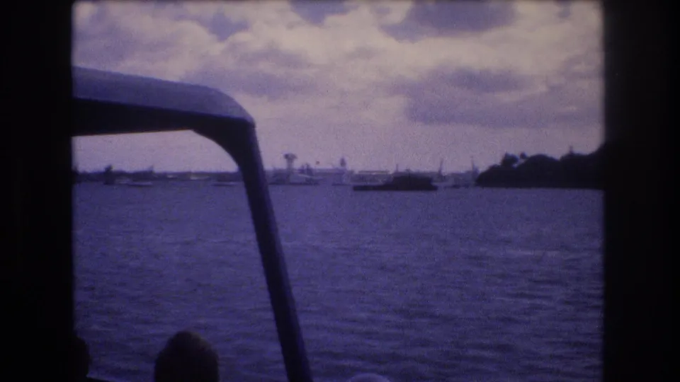 1977 Honolulu boat-view footage with harbor water, several distant boats, a low shoreline, cloudy sky, and dark foreground edges from the vessel or film frame.