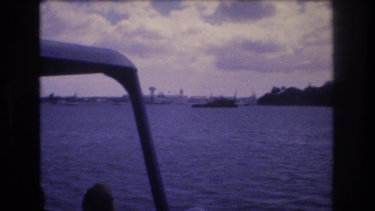 1977 Honolulu home-movie frame from a moving boat, looking past dark canopy supports across rippling water toward a low distant shoreline and small vessels.