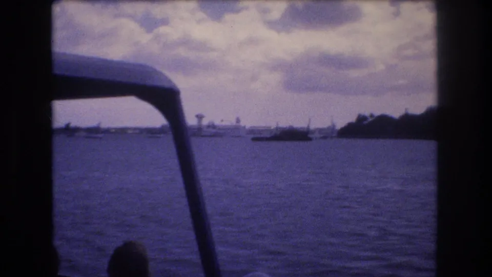 1977 Honolulu home-movie frame from a moving boat, looking past dark canopy supports across rippling water toward a low distant shoreline and small vessels.