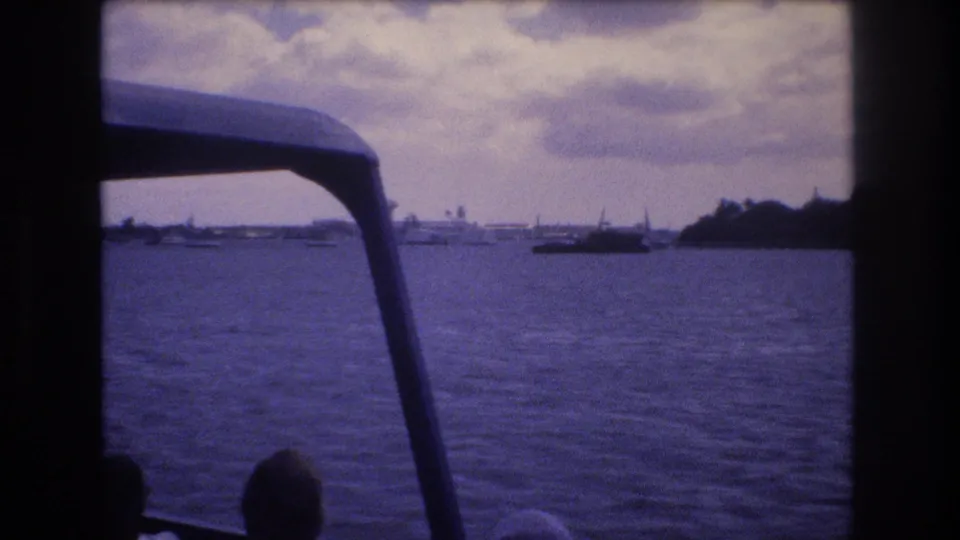 A Honolulu 1977 home-movie view from inside a boat, with foreground rails and canopy supports framing calm harbor water, distant boats, and low shoreline forms.