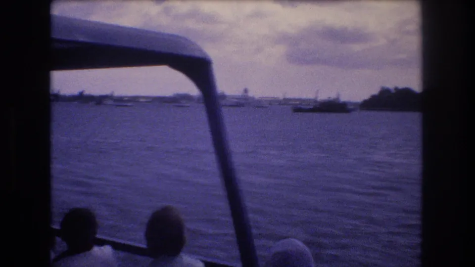 1977 Honolulu visitor-shot boat scene with silhouetted young adults in the foreground, a canopy above, rippled harbor water, and low shoreline shapes in the distance.
