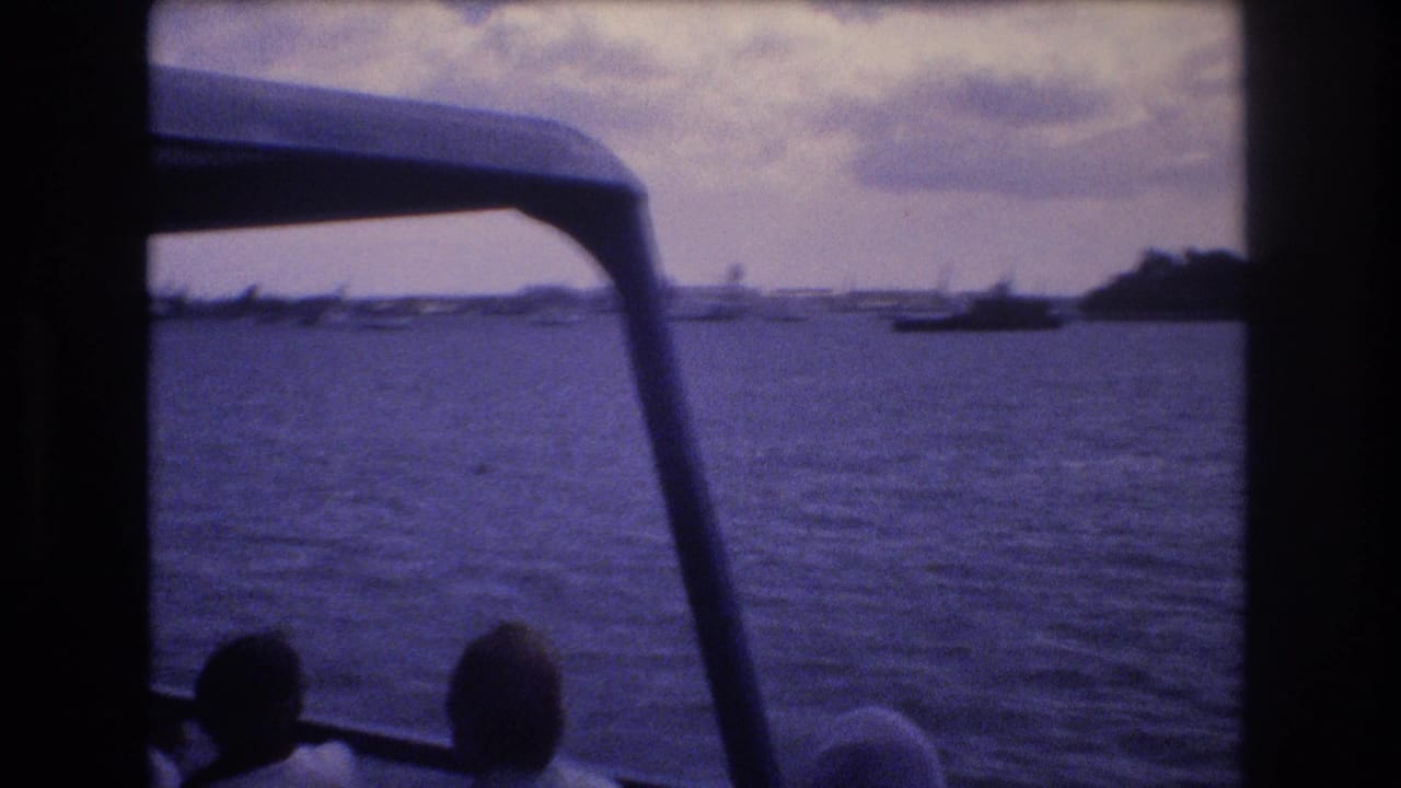 1977 Honolulu home-movie frame looking from inside a moving boat across rippling water toward a distant shoreline and small boats, with passengers silhouetted in the foreground.