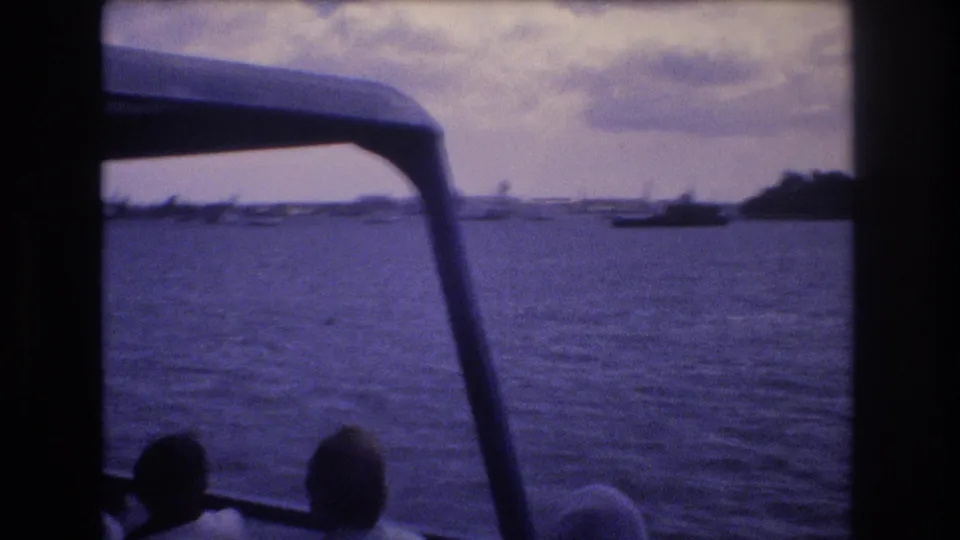 1977 Honolulu home-movie frame looking from inside a moving boat across rippling water toward a distant shoreline and small boats, with passengers silhouetted in the foreground.