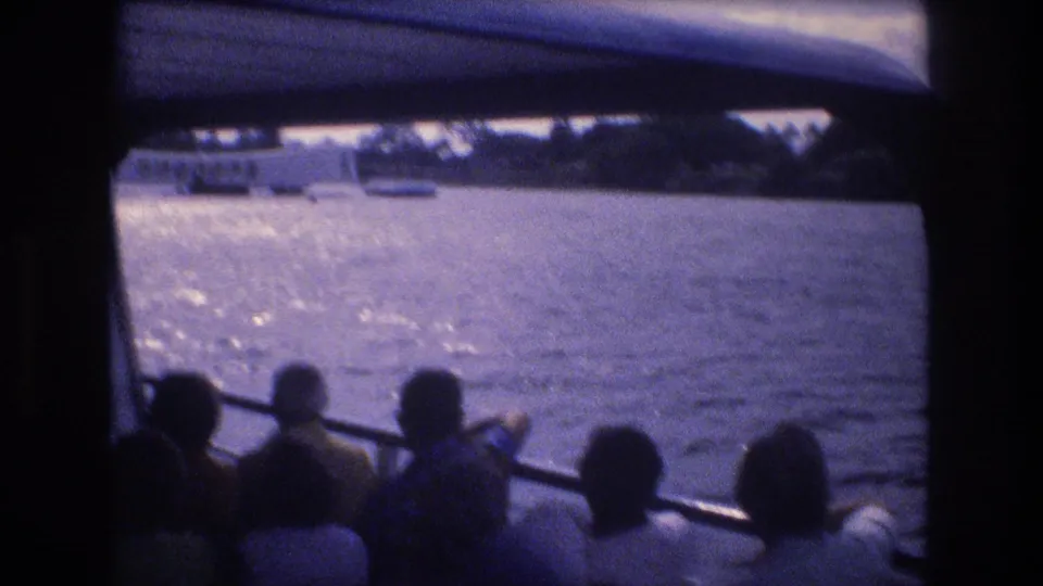 1977 Honolulu home-movie frame with several passengers seated beneath a boat canopy, bright reflections on the water, and small vessels near the distant shore.