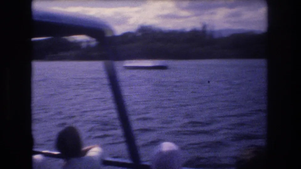 1977 Honolulu boat-ride frame showing passengers at the bottom edge, vertical boat supports, calm water, a small floating object or boat, and a dark distant shoreline.