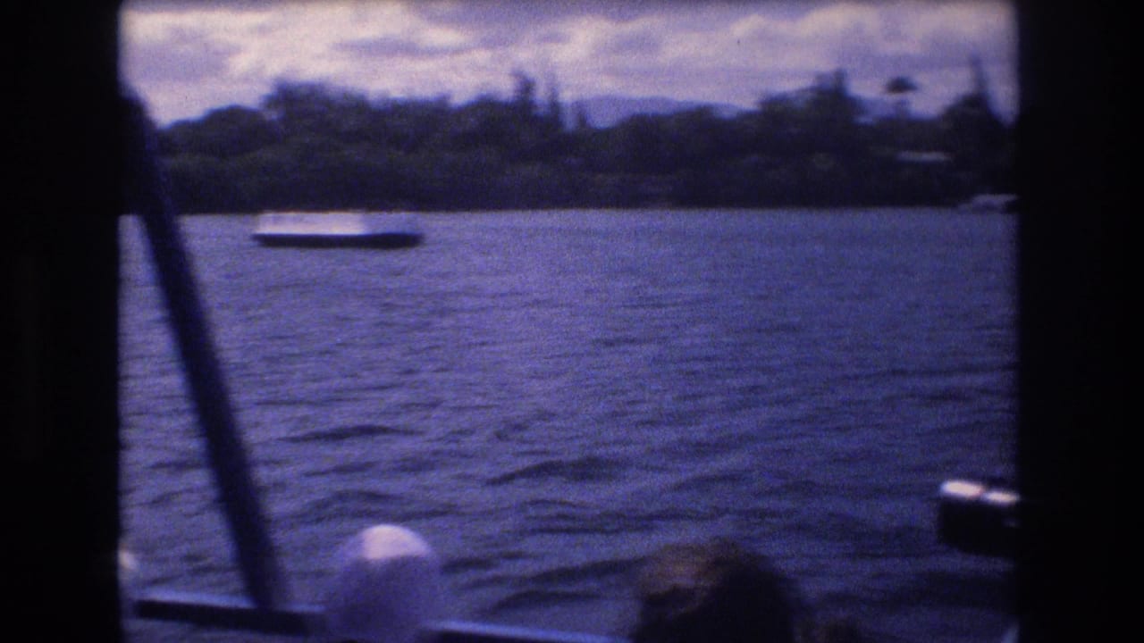 1977 Honolulu home-movie frame looking from inside a boat toward blue harbor water, a low white vessel, cloudy sky, and a green shoreline.