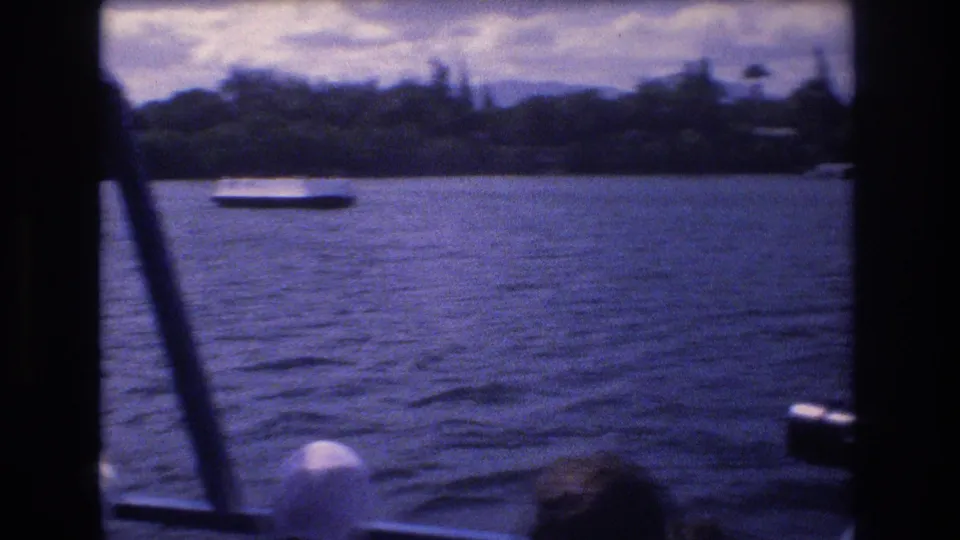 1977 Honolulu home-movie frame looking from inside a boat toward blue harbor water, a low white vessel, cloudy sky, and a green shoreline.