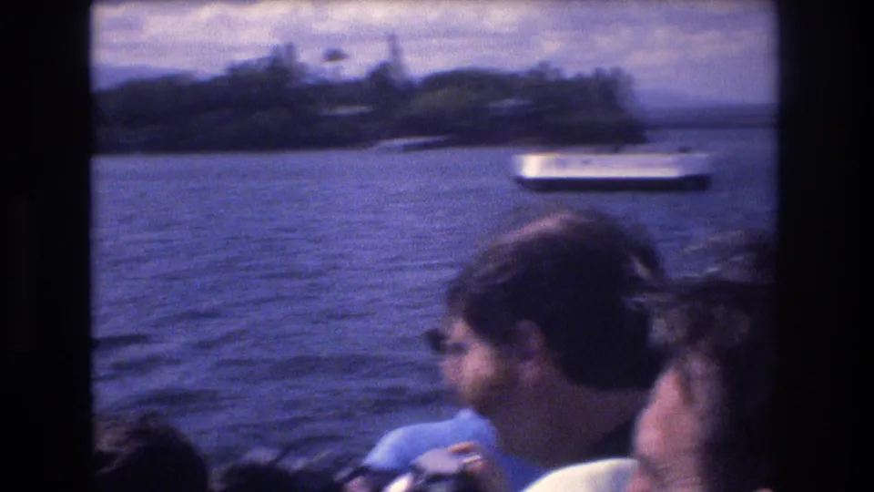 Passengers aboard a boat in Honolulu look out toward blue water, a distant white passenger boat, and a tree-lined shoreline in 1977 home-movie footage.