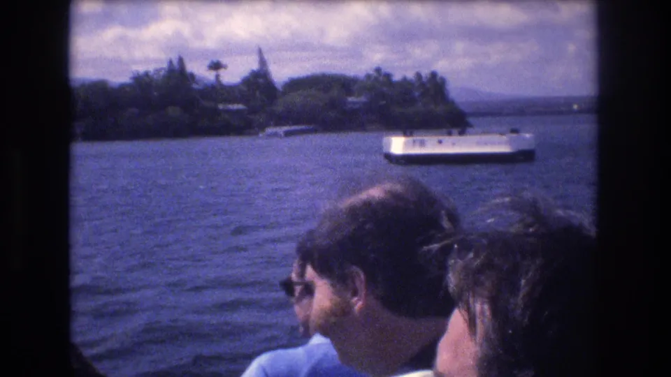 A man in glasses and other passengers are seen from the boat interior while a white ferry or tour boat crosses the water near a palm-lined Honolulu shore.