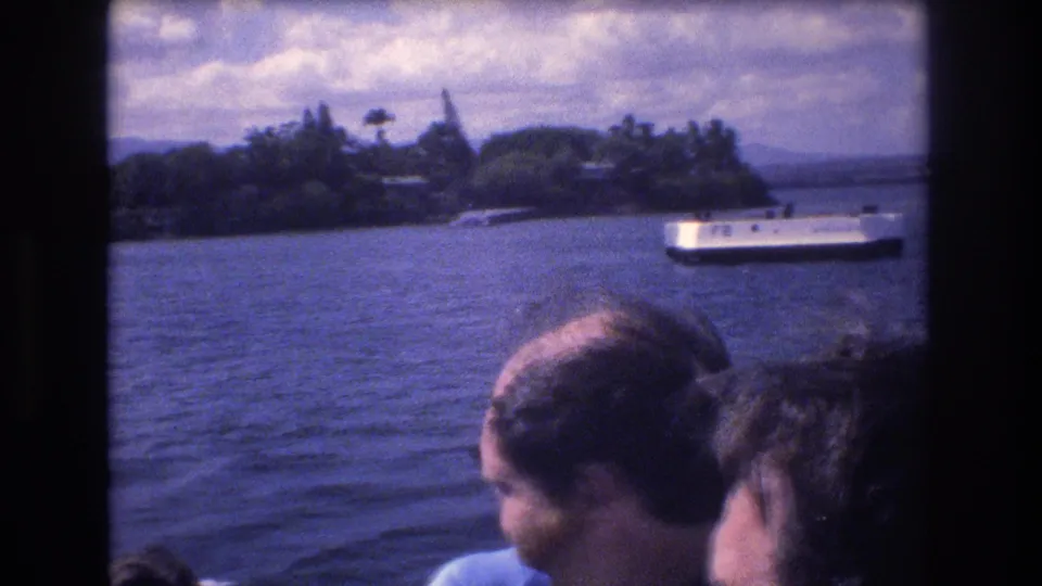 Late-frame view from inside a Honolulu boat with passengers in the foreground, blue harbor water, a white passenger boat, and green shoreline under clouds.