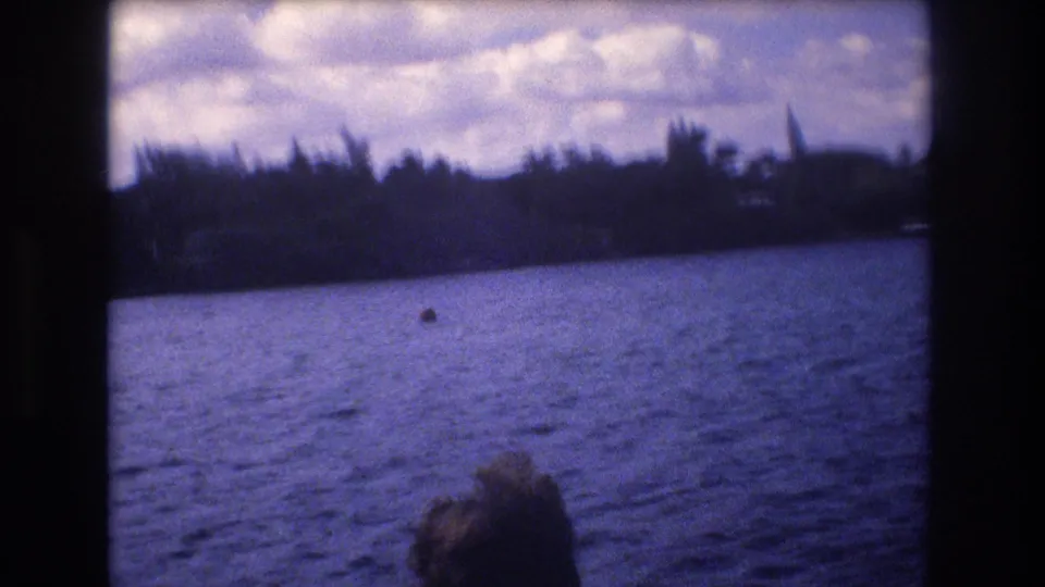 A handheld 8mm view from a boat shows blue water, a distant tree-lined Honolulu shore, clouds, and a passenger's head at the bottom edge.